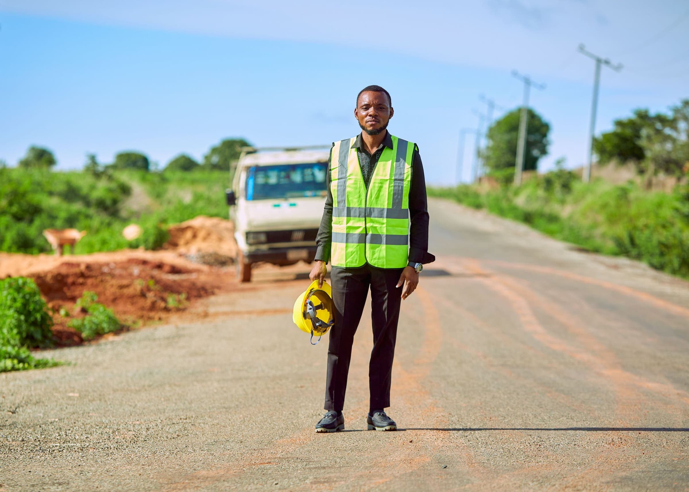 Un ingénieur en gilet haute visibilité, casque à la main, debout sur une route rurale à côté d’un camion à l’arrêt.