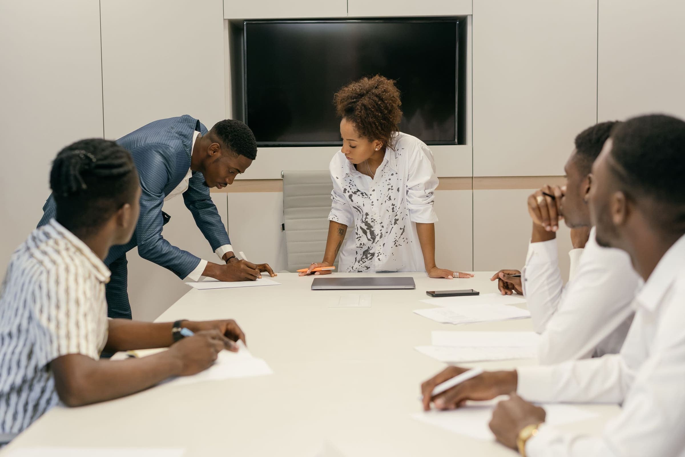 Une femme debout en bout de table de réunion, pendant que ses collègues examinent des documents et prennent des notes.
