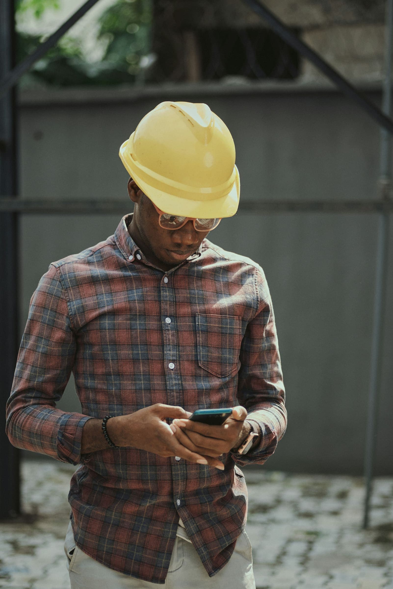 Un ingénieur de chantier en casque jaune et lunettes de sécurité consulte son téléphone à l’extérieur.