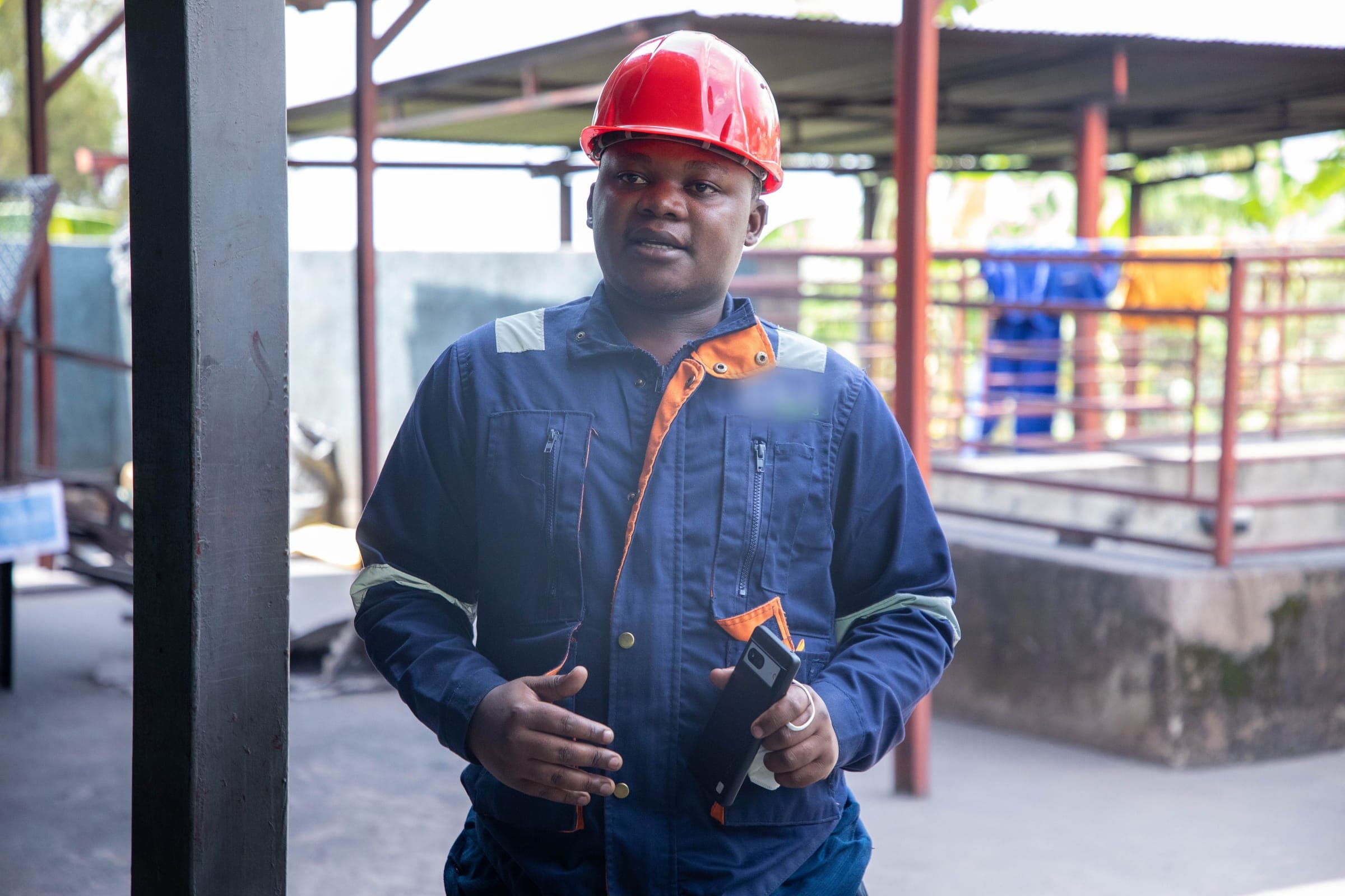 Un superviseur en casque rouge et combinaison bleu marine, téléphone à la main, dans une usine de traitement de minerai.